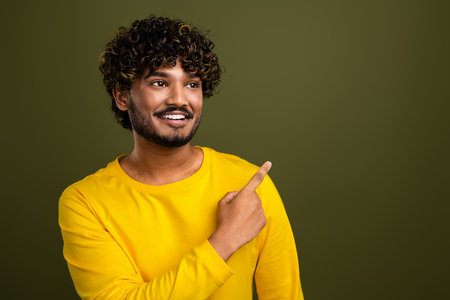 Smiling young man wearing a yellow shirt pointing excitedly to the side with a cheerful expression against a green backgroundの写真素材