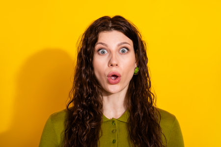Surprised young woman with curly brown hair on a vibrant yellow background wearing fashionable green apparelの写真素材