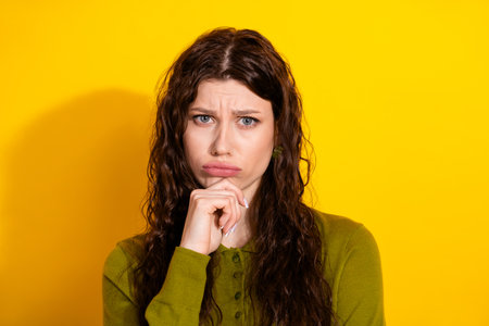 Portrait of a thoughtful young woman in a green outfit against a yellow background, expressing contemplation with hand gestureの写真素材
