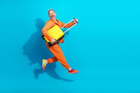 Smiling man in orange suit holding yellow suitcase, mid leap against a vibrant blue background for...