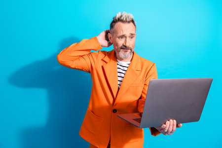Middle aged man with blonde dyed hair in an orange blazer, holding a laptop and showing an uncertainの写真素材