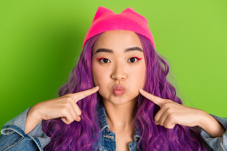 Stylish young woman with dyed purple hair posing on green background wearing pink beanie and denim...