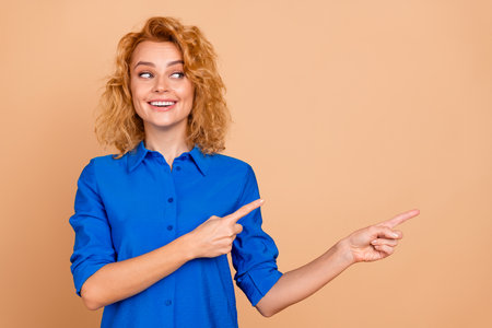 Young businesswoman in blue shirt gesturing with a cheerful smile, promoting concepts of positivity and success on beige backgroundの写真素材