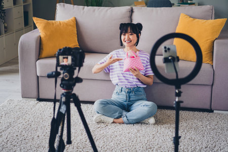 Photo of shiny cute girl dressed purple striped t-shirt recording vlog modern gadget showing cosmetics bag indoors house apartment roomの写真素材
