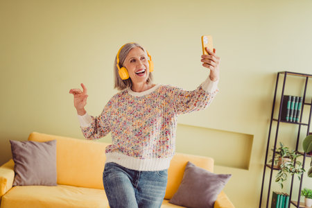 Elderly woman joyfully taking a selfie indoors with headphones, wearing a stylish knitted pullover