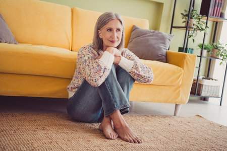 Charming elderly woman enjoying a leisurely day at home in a cozy living room with a bright yellow sofa and stylish sweaterの写真素材