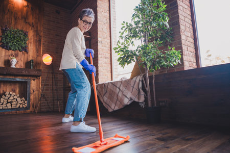Charming elderly woman enjoying a leisurely weekend at home, cleaning her modern apartment with joy...