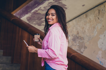 Photo of attractive young woman lifting stairs hold device coffee paper cup wear pink clothes office building interior indoors workspaceの写真素材