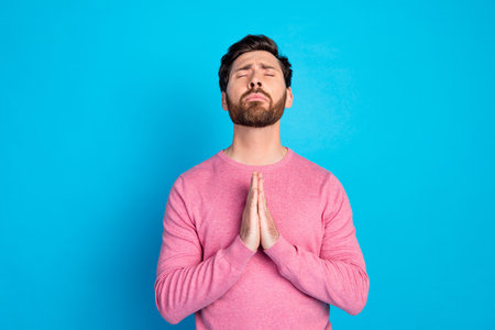 Peaceful man in casual pink sweater posing with prayer gesture against vibrant blue backgroundの写真素材