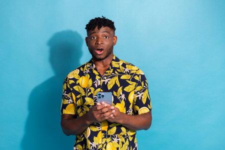 Stylish man with surprised expression in trendy shirt against blue background, holding smartphone,...