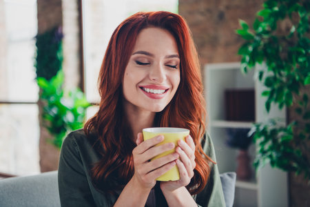 Photo of lovely young ginger girl hold coffee mug smell aroma dressed khaki garment spacious house indoors roomの写真素材