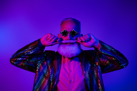 Photo of attractive funny senior guy wear shiny jacket star glasses enjoying party touching mustache isolated violet neon color backgroundの写真素材