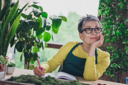 Photo of pensioner lady florist look dreamy empty space write notebook wear green apron working flower shop studio small businessの写真素材