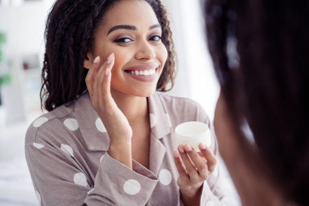 Young woman with afro hair smiling while applying face cream in cozy pajamas in morning lightの写真素材