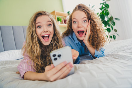 Full size photo of two young women excited hold device lying bed dressed sleepwear outfit wake up cozy day light bedroom home interiorの写真素材