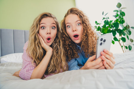 Full size photo of two attractive young women amazed hold device lying bed dressed sleepwear outfit wake up day light bedroom home interiorの写真素材