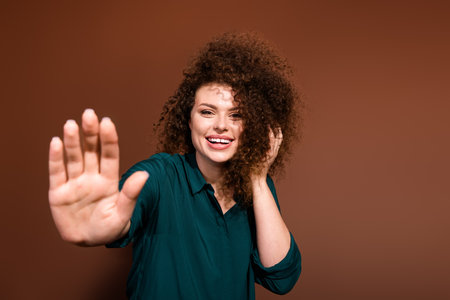 Young woman with curly brunette hair in a green shirt smiles against a brown background while...