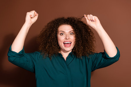 Joyful young woman celebrating with curly hair and green shirt on brown backgroundの写真素材