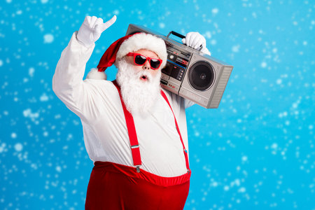 Photo of cool hispter santa claus with beard stomach listen x-mas christmas boom box wear red style stylish trendy suspenders overalls cap isolated over blue color backgroundの写真素材