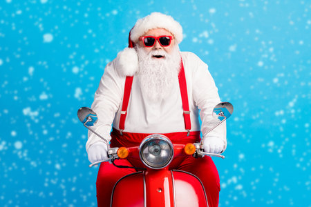 Photo of grandpa driver grey beard ride fast retro motorbike smile wear santa claus x-mas costume suspenders sunglass white shirt headwear gloves isolated blue color backgroundの写真素材