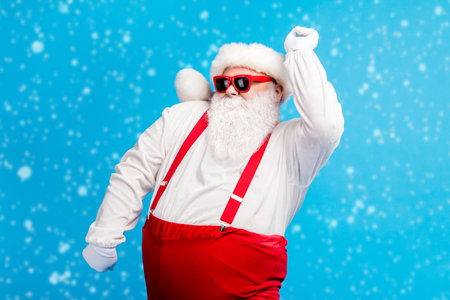 Photo of funky old man grey beard dance nightclub raise hand wear santa claus x-mas costume cap headwear sunglass suspenders white shirt gloves isolated blue color backgroundの写真素材