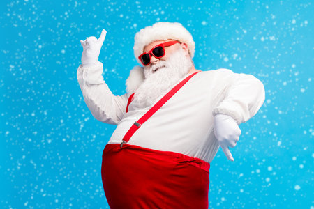 Photo of old man grey beard dance direct fingers raise hand wear santa claus x-mas costume cap headwear sunglass suspenders white shirt gloves isolated blue color backgroundの写真素材