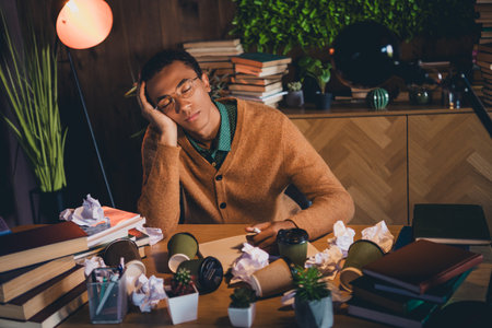 Young student falling asleep while studying at home. African male in casual jumper surrounded by books and notes in evening.の写真素材