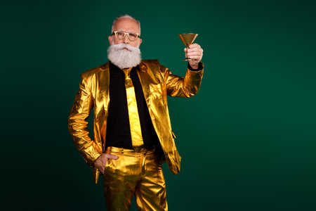 Elegant elderly man in golden suit celebrating with a drink against a vibrant green backdrop, perfect for holiday and celebration themesの写真素材
