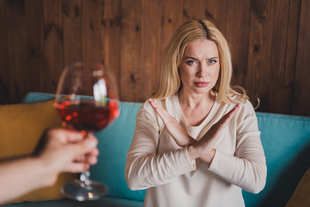 Photo of confident serious woman dressed beige shirt showing stop gesture drinking alcohol indoors house apartment roomの写真素材