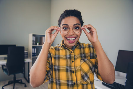 Photo of attractive young woman touch spectacles excited wear yellow plaid shirt comfortable modern office room interior indoors workspaceの写真素材