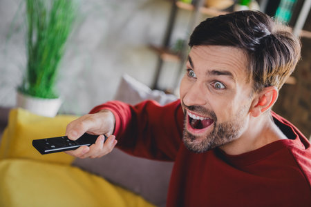 Enthusiastic young man enjoying his weekend indoors, immersed in a riveting show, wearing a cozy...