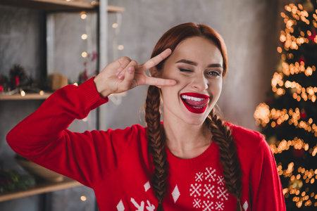 Photo of pretty young girl v-sign eye wink lick lips wear sweater christmas festive holiday time apartment indoorsの写真素材