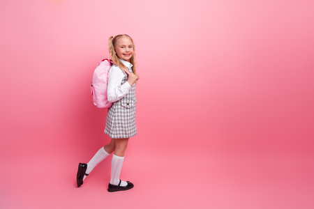 Smiling young schoolgirl in fashionable uniform carrying a pink backpack on a bright pink background, representing youth and educationの写真素材