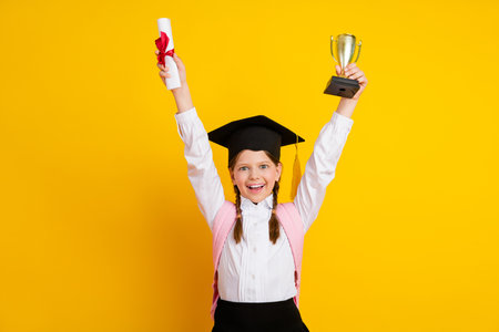 Happy schoolgirl holding a diploma and trophy during academic achievement celebration on yellow backdropの写真素材