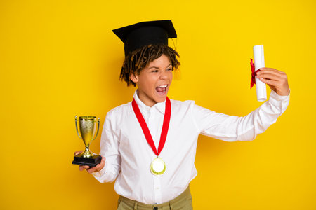 Joyful schoolboy celebrates educational success holding diploma and trophy on vibrant yellow backgroundの写真素材