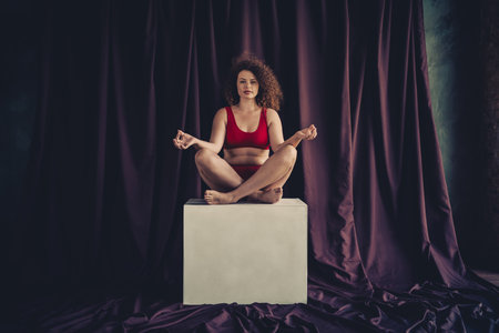 Young woman with curly hair sitting confidently on a cube in a studio with a purple textile backdrop wearing red lingerieの写真素材