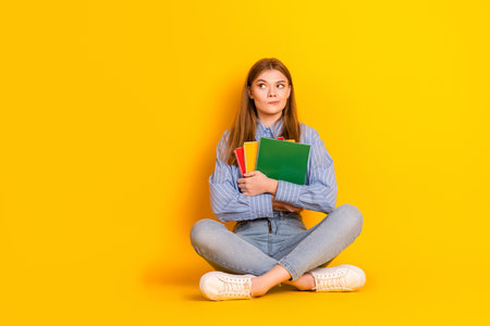 Young woman sitting cross-legged holding notebooks on a yellow background showcasing casual lifestyle and education themeの写真素材