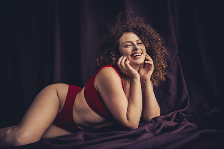 Confident young woman embracing natural beauty in stylish red lingerie against a purple textile studio backdropの写真素材