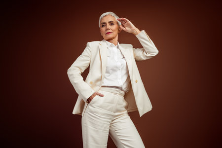 Confident mature businesswoman in an elegant white suit posing against a brown backgroundの写真素材