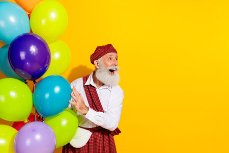 Cheerful senior Scottish man in traditional clothing holding vibrant balloons on yellow background...