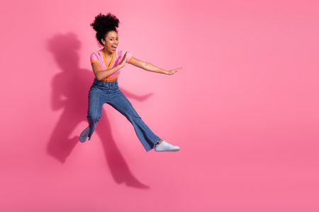 Cheerful young woman jumping in stylish casual attire expressive against pink background, expressing...