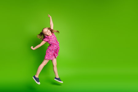 Happy young girl in vibrant pink dress jumping against a bright green background showcasing joy and playfulnessの写真素材