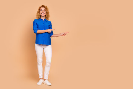 Cheerful young woman in stylish casual attire pointing aside on beige background for promotion or advertising conceptsの写真素材