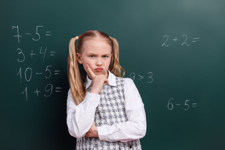 Thoughtful schoolgirl in uniform ponders math problems on chalkboard during classroom lesson in...