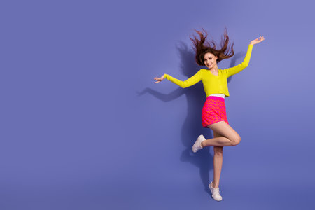 Cheerful young woman jumping energetically in stylish outfit against purple background with vibrant...