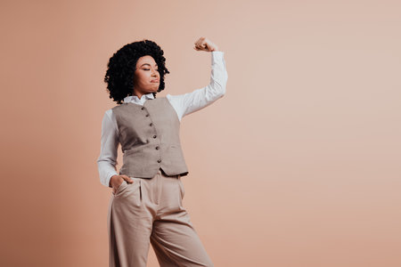Confident businesswoman in formal attire flexing an arm on a beige background showcasing strength, professionalism, and styleの写真素材