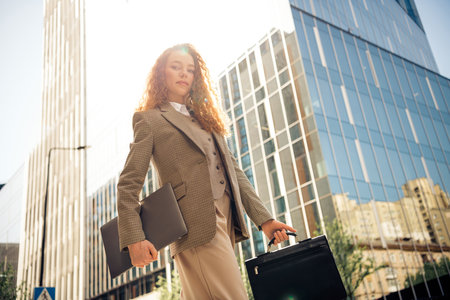 Confident businesswoman with laptop and briefcase standing outdoors in urban city setting on a bright sunny dayの写真素材