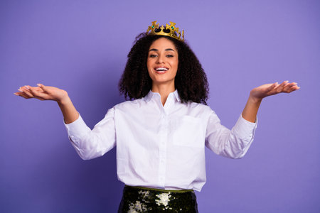 Confident young woman wearing a crown and stylish attire smiling against a purple background with extended armsの写真素材