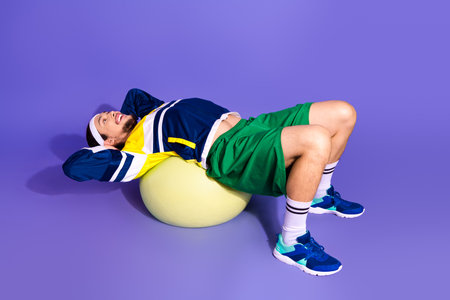 Active Sportsman Relaxing on Exercise Ball While Wearing Retro Style Activewear Against Purple Backgroundの写真素材