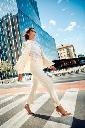 Confident businesswoman walking outdoors in formal wear near modern office building in a dynamic sunny city street.の写真素材
